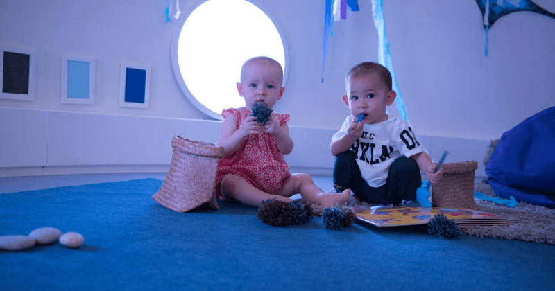 Baby sitting on a blue cushioned floor, reaching out to hold a soft toy, in a calm, sensory-rich learning space.
